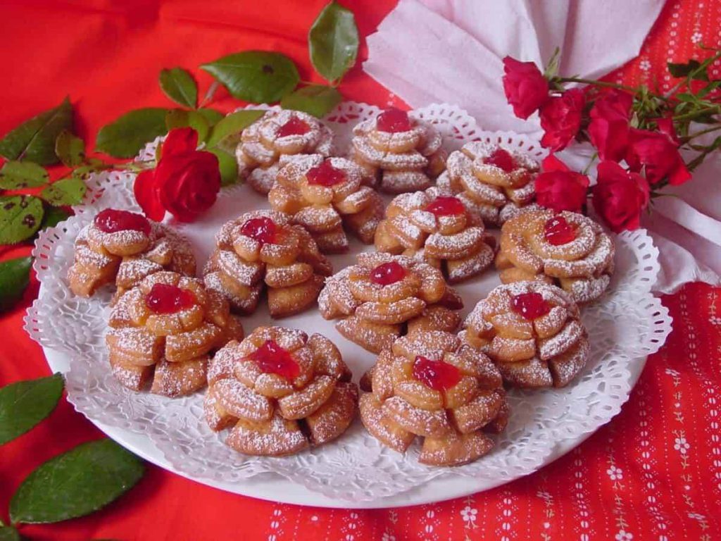 Rosenkrapfen auf einem Teller, dekoriert mit Puderzucker und Kirschen, umgeben von Rosen.