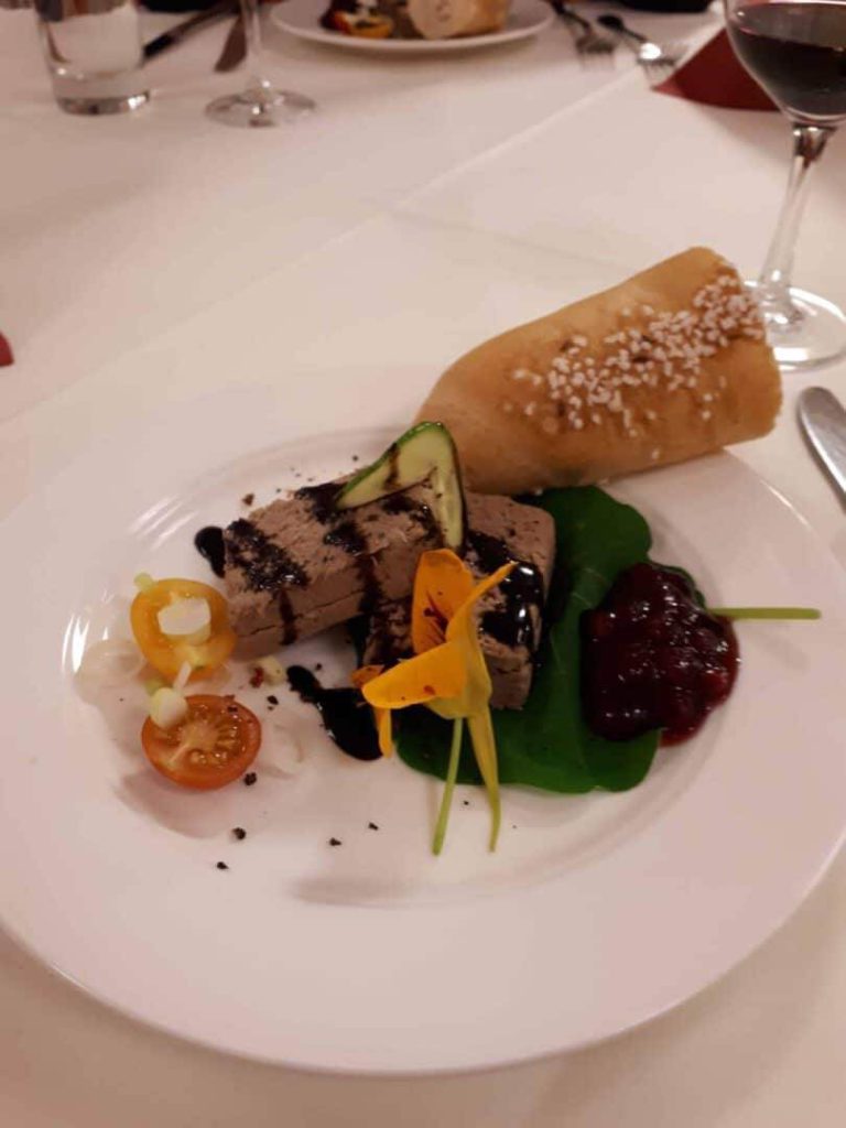 Wildfleischpastete mit Garnitur aus Gemüse, Blüten und einer Beilage aus Brot.