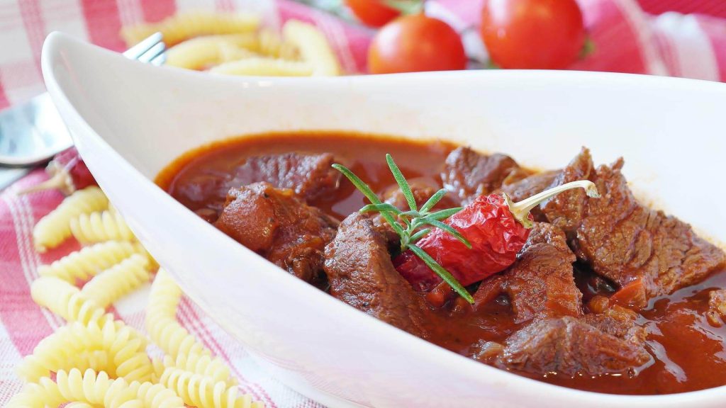 Rindsgulasch in einer weißen Schüssel, umgeben von frischen Zutaten und Nudeln.
